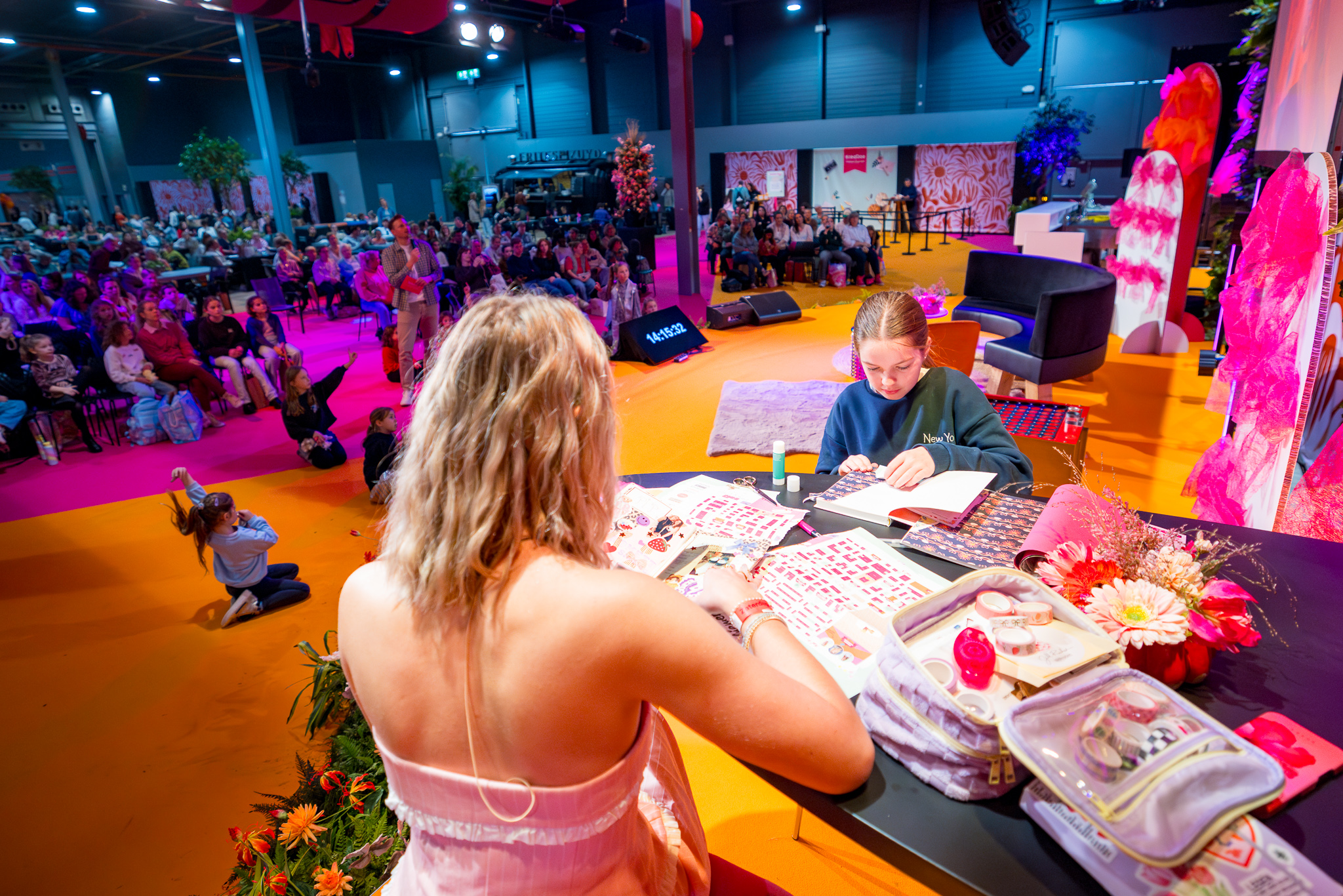 Presentatrice en meisje plakken stickers in knutselboek aan tafel op podium, terwijl publiek in zaal toekijkt
