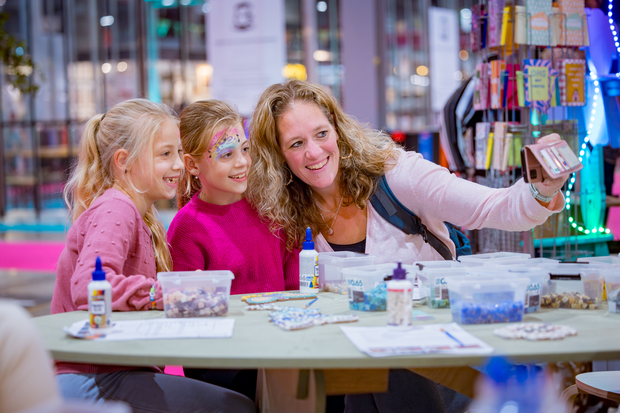 Moeder maakt een selfie met twee meisjes aan een knutseltafel vol bakjes met blauwe, witte en bruine kralen en flesjes lijm.