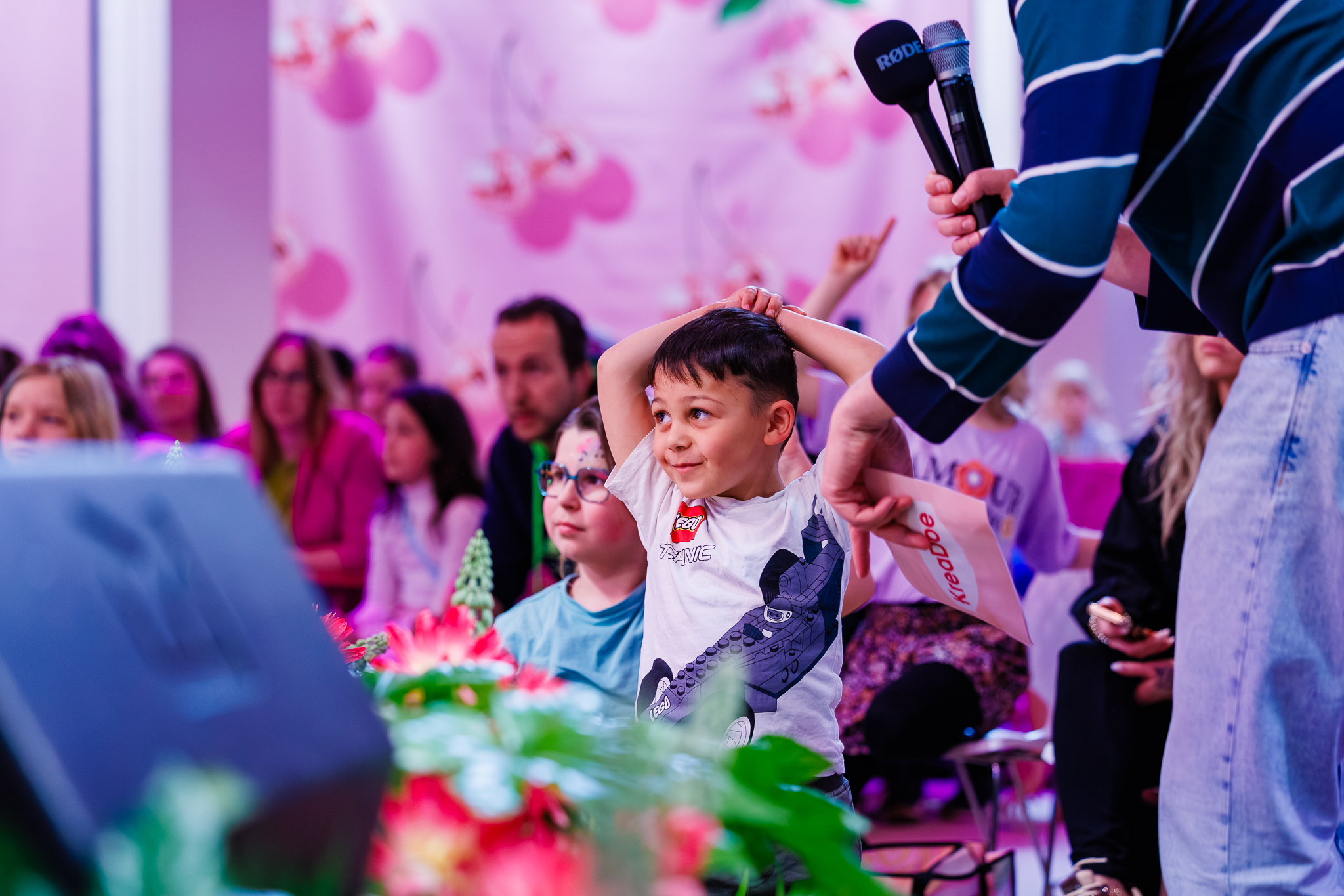 Jongen in wit T-shirt met Lego-print steekt zijn armen boven zijn hoofd tijdens een quiz of spel, terwijl een presentator met een zwarte microfoon en een wit kaartje vragen stelt aan het publiek.