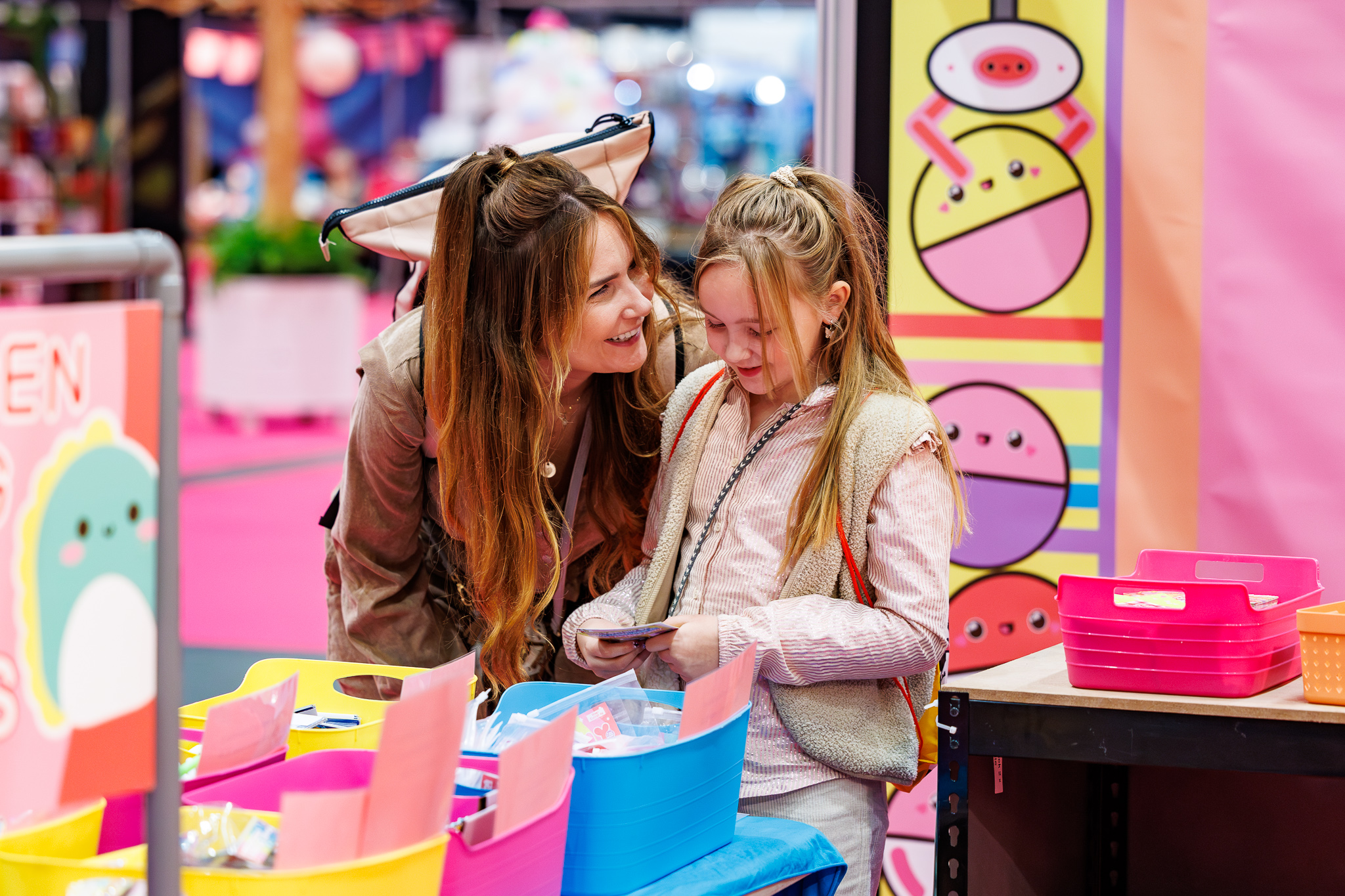 Moeder buigt lachend naar meisje dat kaartjes bekijkt bij bakken met roze, gele en blauwe knutselspullen op een creatieve beurs.