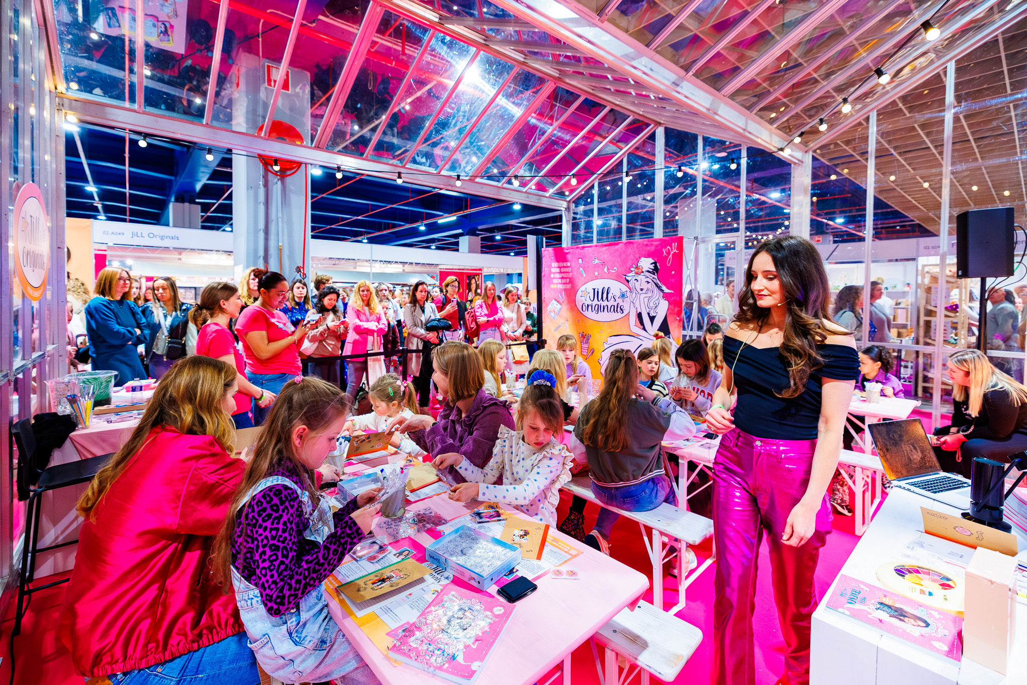 Kinderen knutselen aan lange tafels met stickers en kaartjes tijdens workshop, terwijl presentatrice in zwarte top en roze broek rondloopt.