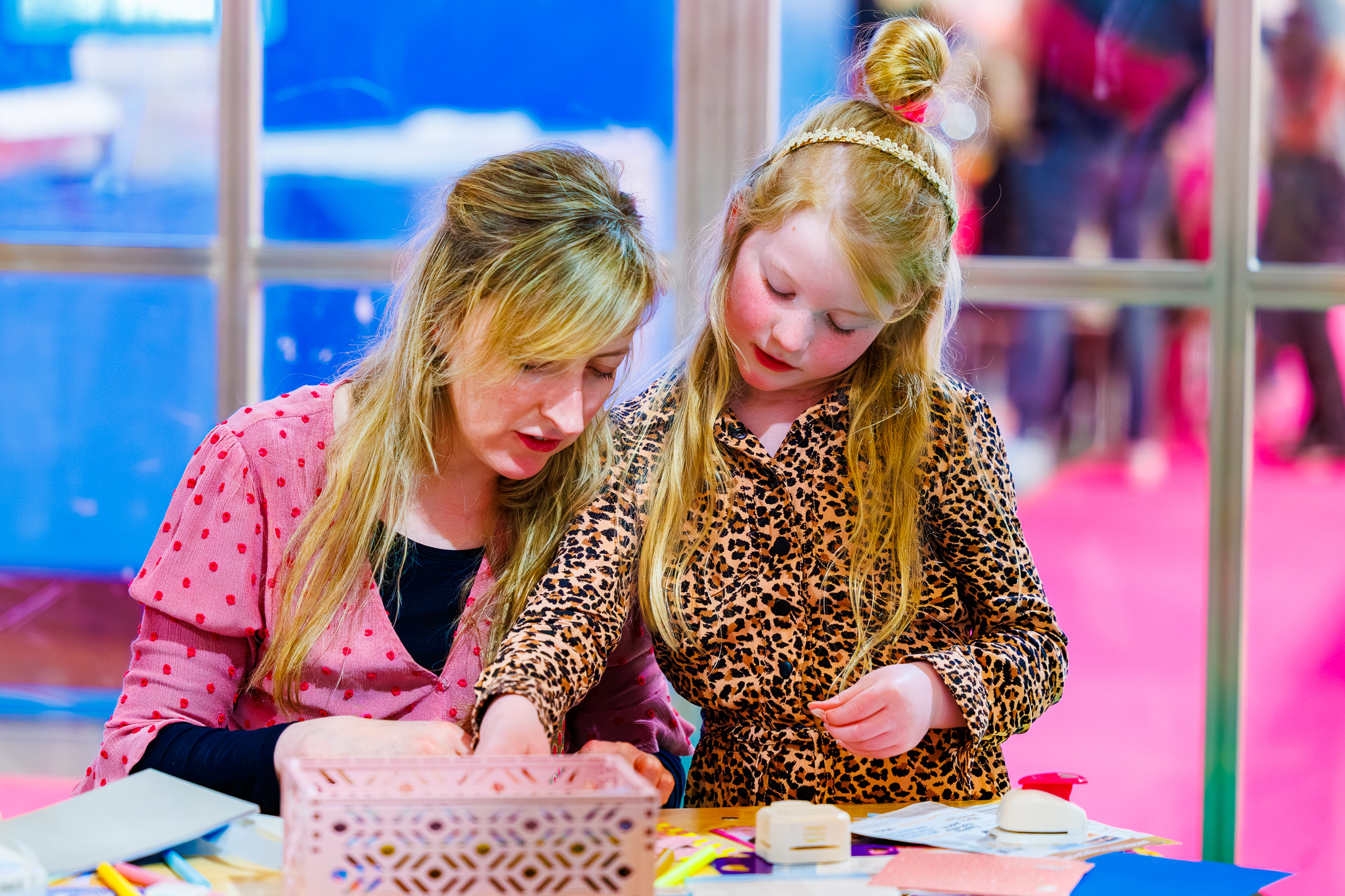 Moeder in roze blouse en meisje in luipaardjurk pakken knutselmateriaal uit een roze bakje aan een tafel.