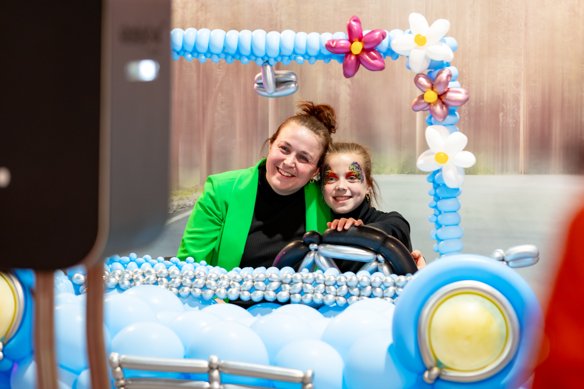 Vrouw en meisje met schmink poseren lachend in blauwe ballonauto met bloemen, terwijl een foto van hen wordt gemaakt.